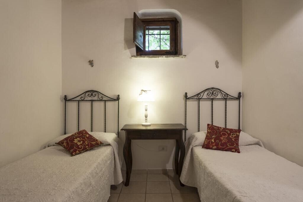 Ein kleiner, schwach beleuchteter Raum mit zwei Einzelbetten, einem kleinen Tisch und einem Fenster mit Blick auf Grünfläche.