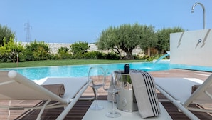 Un espace aménagé au bord de la piscine, avec des chaises longues, une table avec des verres à vin et une bouteille, et une vue sur la piscine et la végétation.