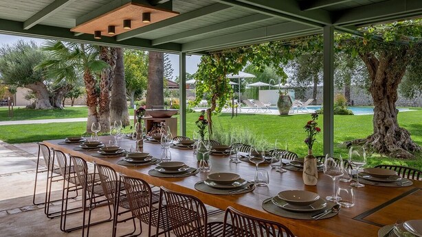 Un espace de restauration en plein air avec une longue table dressée pour un repas, entourée de chaises, et offrant une vue sur une piscine et un espace verdoyant.