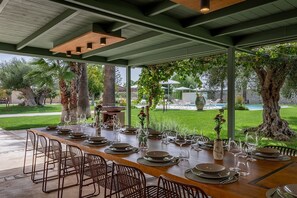 An outdoor dining area with a long table set for a meal, surrounded by chairs, with a view of a pool and greenery. - Villa Aulina, Private villa near Modica (Modica)