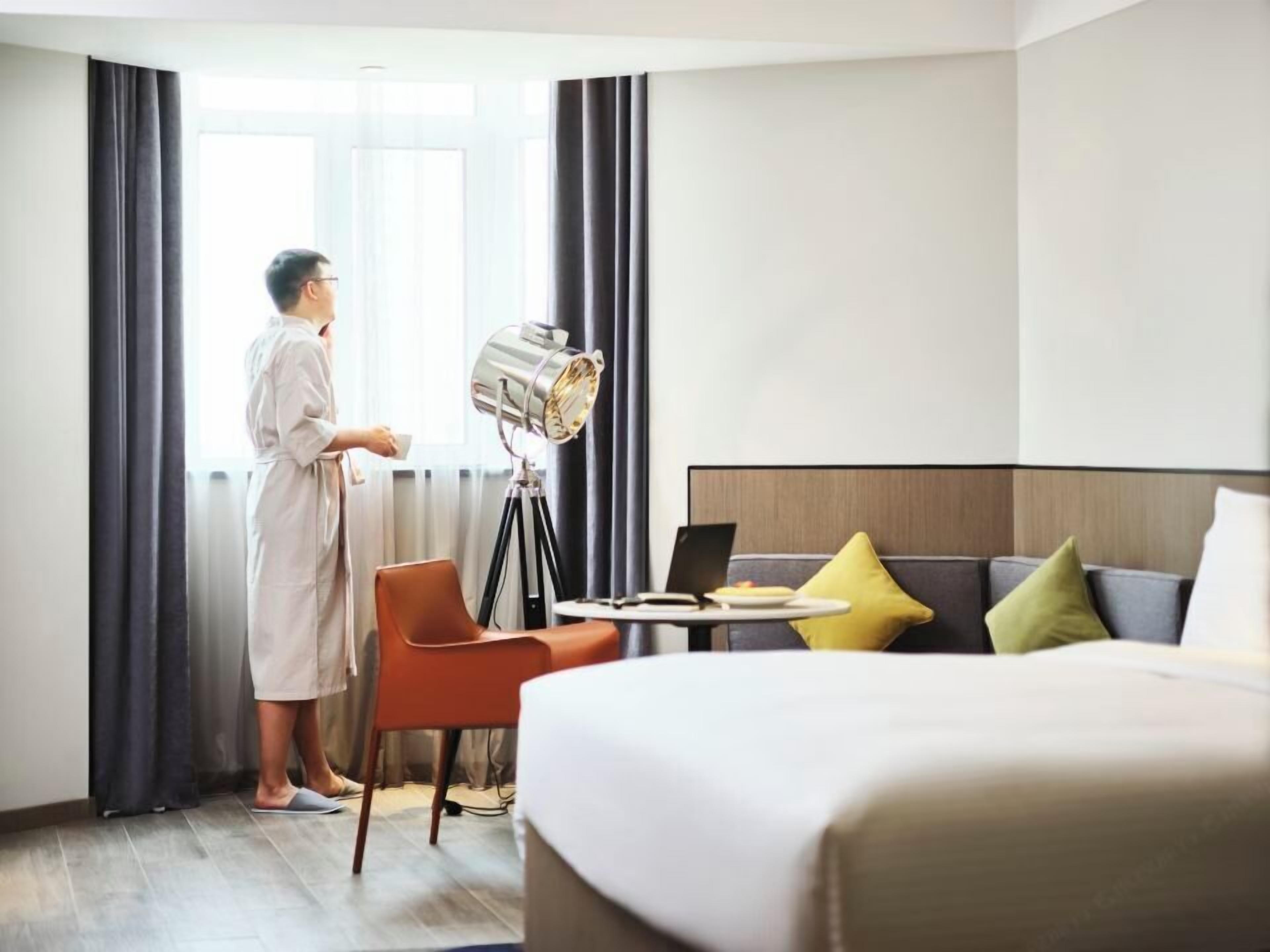 Ein Hotelzimmer mit Bett, Schreibtisch mit Laptop, Stuhl und großem Fenster mit Vorhängen.