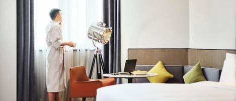 Habitación de hotel con cama, escritorio con laptop, silla y ventana grande con cortinas.