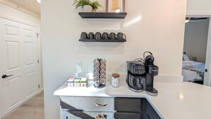 Une cuisine compacte équipée d’une machine à café, d’une bouilloire et d’une étagère où sont rangées des tasses et une citation inspirante encadrée.