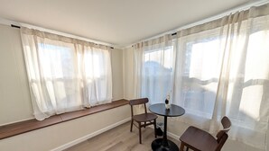 Une pièce dotée d’un plancher en bois, d’une table ronde avec un pied noir, de deux chaises en bois, d’un banc placé le long de la fenêtre et de rideaux légers aux fenêtres.