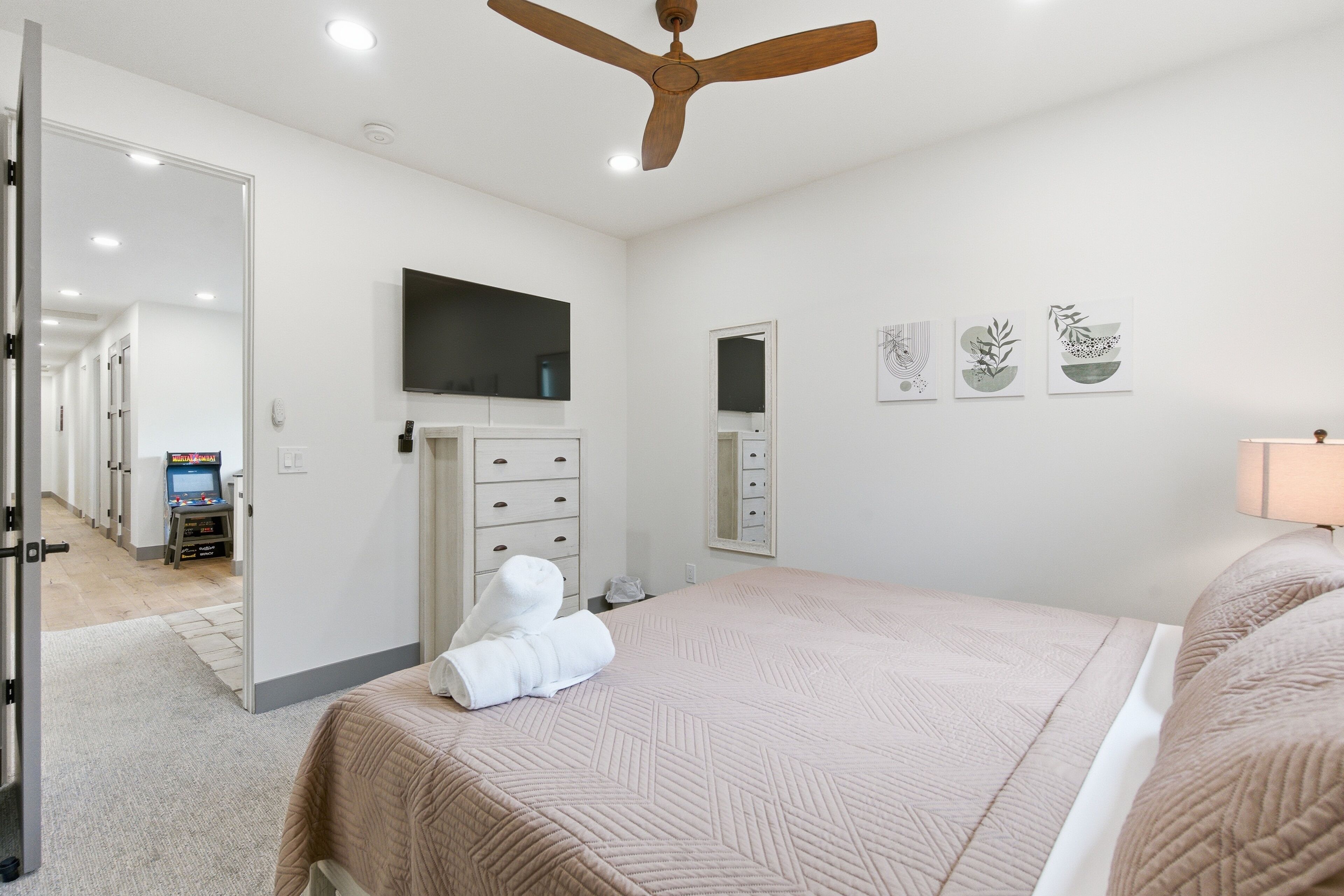 A hotel room with a large bed, a ceiling fan, a flat-screen TV, a minibar, and artwork on the walls.