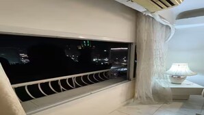 Een balkon met uitzicht op een stadsbeeld 's nachts, een witte lamp op een nachtkastje en een wit gordijn.