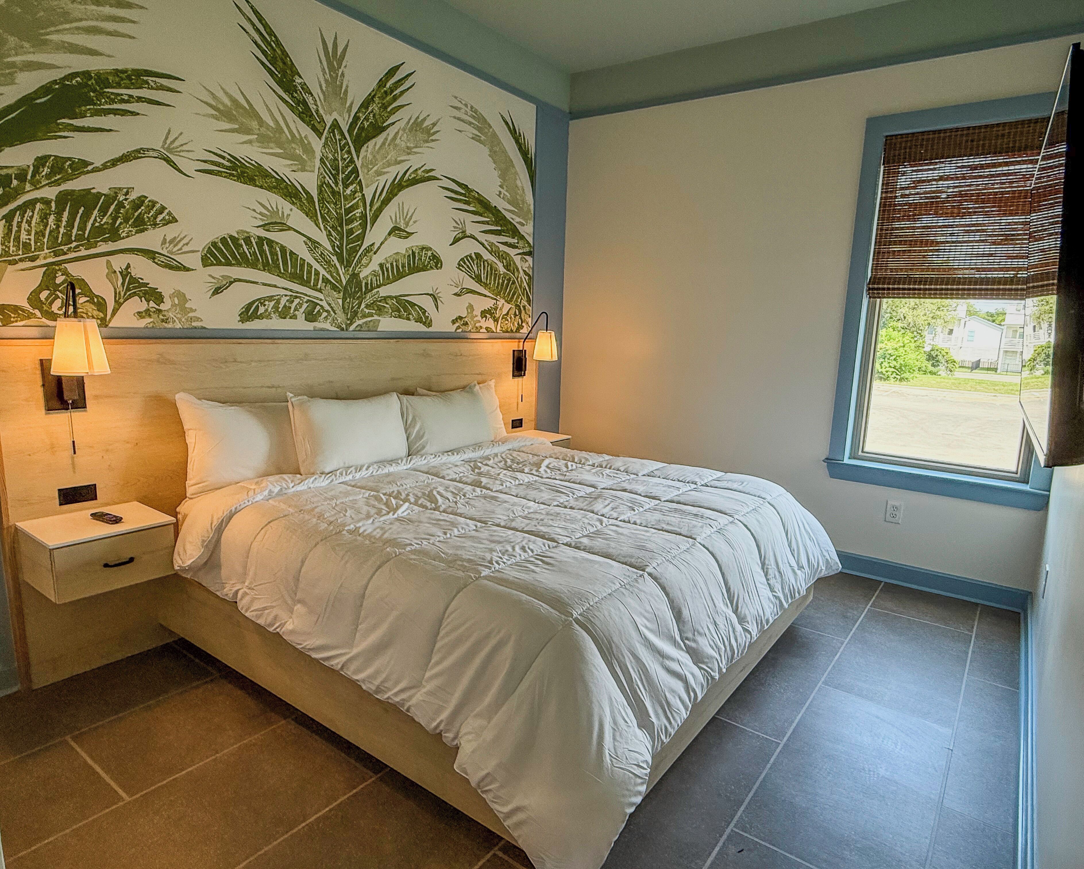 A bedroom with a bed, nightstands, a wall mural, a window with blinds, and a view of the outside.