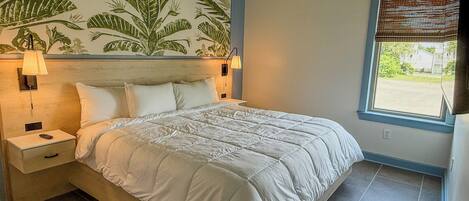 A bedroom with a bed, nightstands, a wall mural, a window with blinds, and a view of the outside.