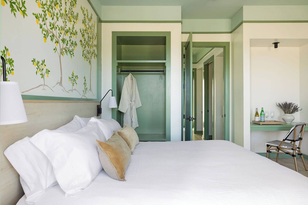 A hotel room with a bed, a wall-mounted lamp, a towel rack, a chair, and a small table with bottles and glasses.