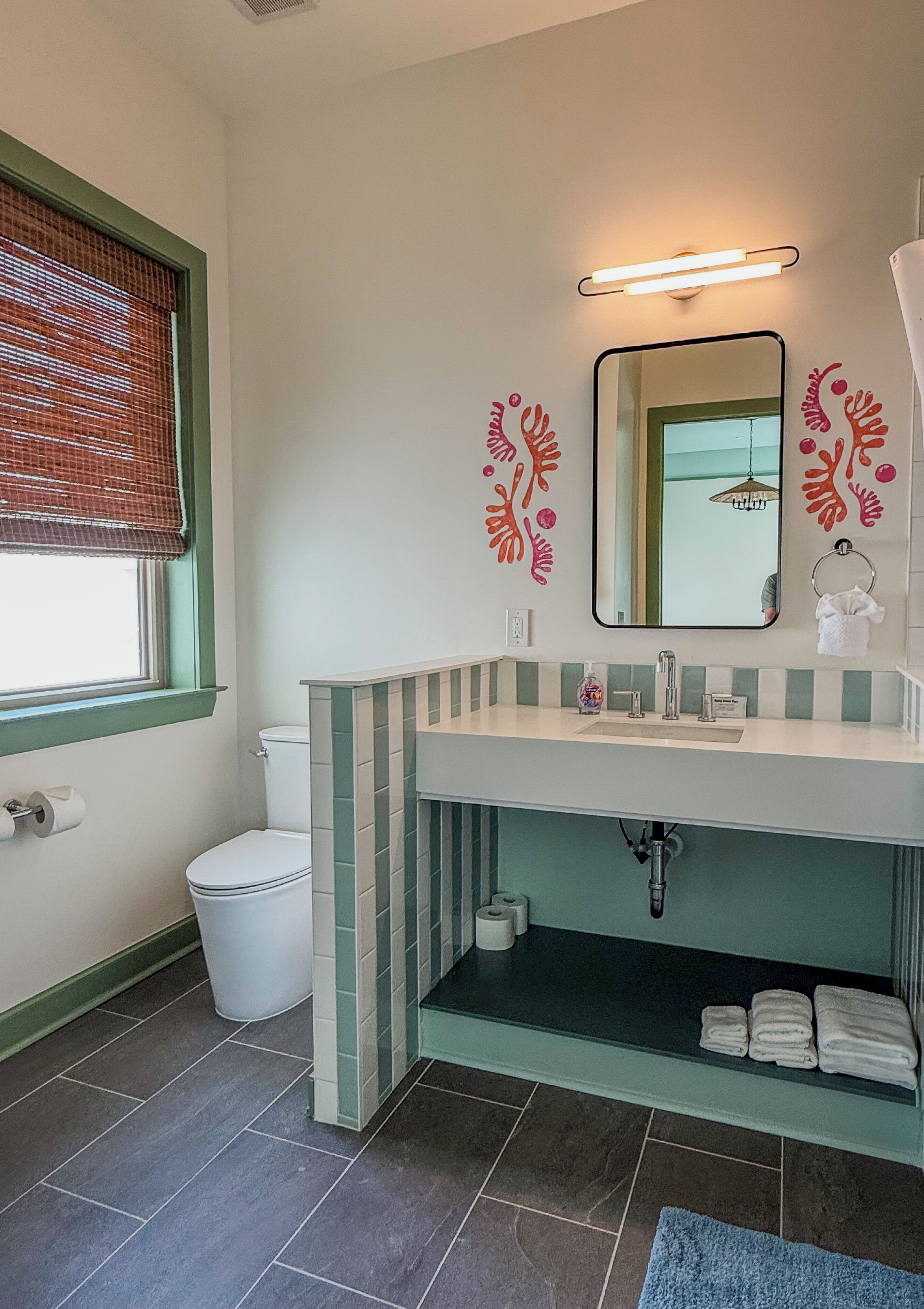 Une salle de bain comprenant des toilettes, un lavabo avec un miroir, un porte-serviettes et une fenêtre avec des stores.