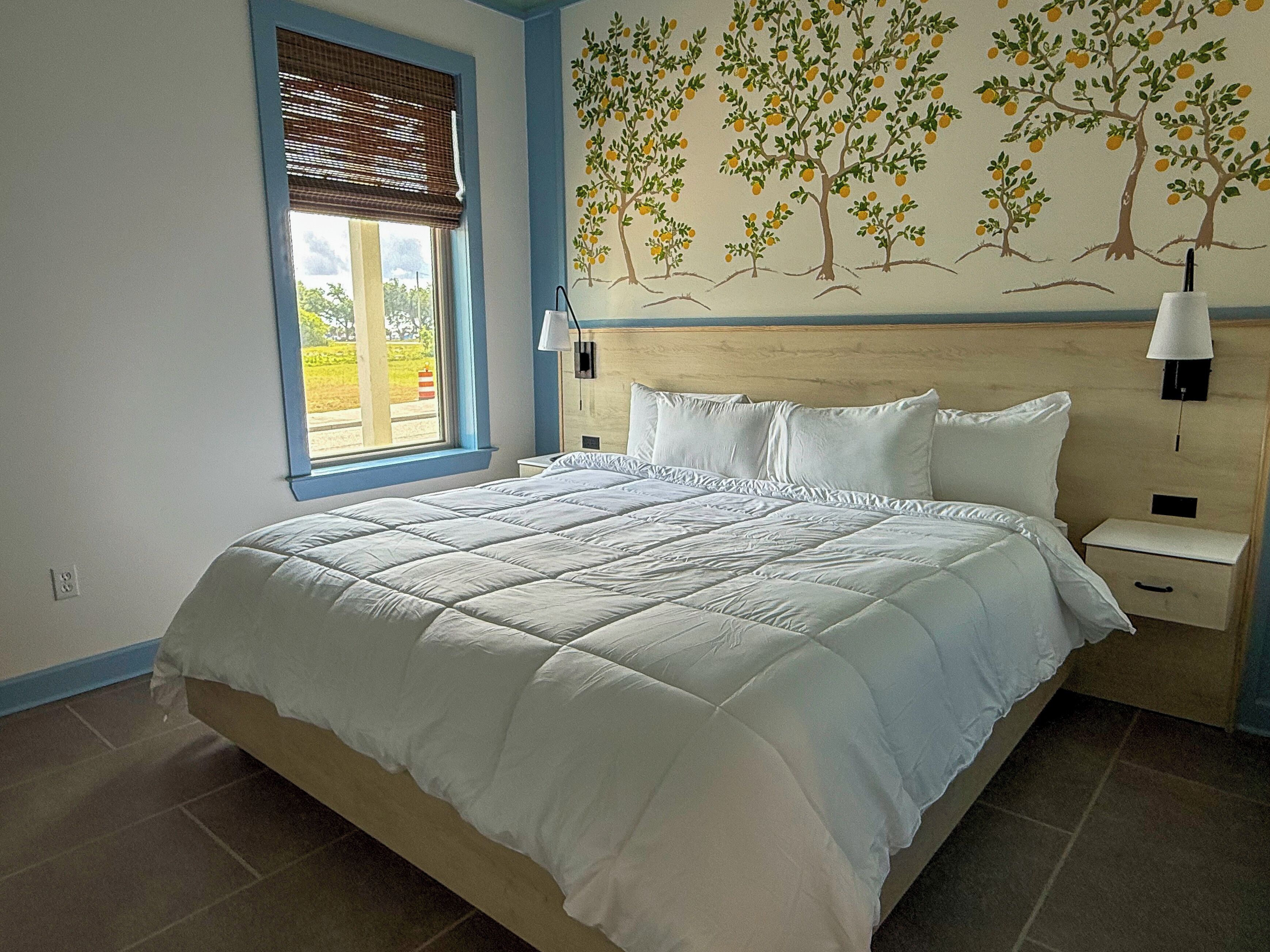 A bedroom with a large bed, a window with blinds, a nightstand, a lamp, and a mural of trees on the wall.