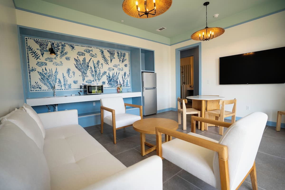 A modern living room with a sofa, chairs, a round table, a kitchen area with a refrigerator, and a wall-mounted TV.