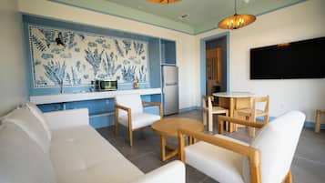 A modern living room with a sofa, chairs, a round table, a kitchen area with a refrigerator, and a wall-mounted TV.