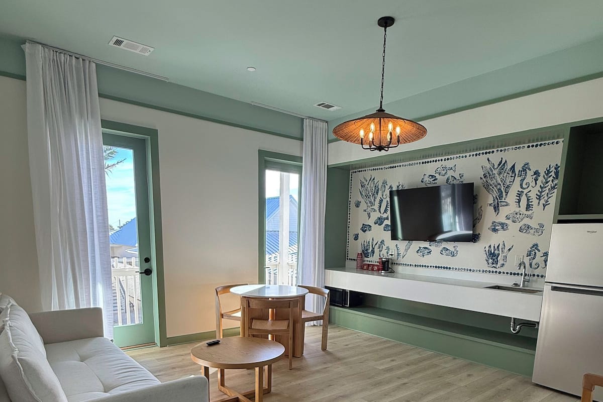 A living room with a sofa, a dining table with chairs, a TV mounted on a wall with a decorative backsplash, and a kitchen area with a refrigerator.