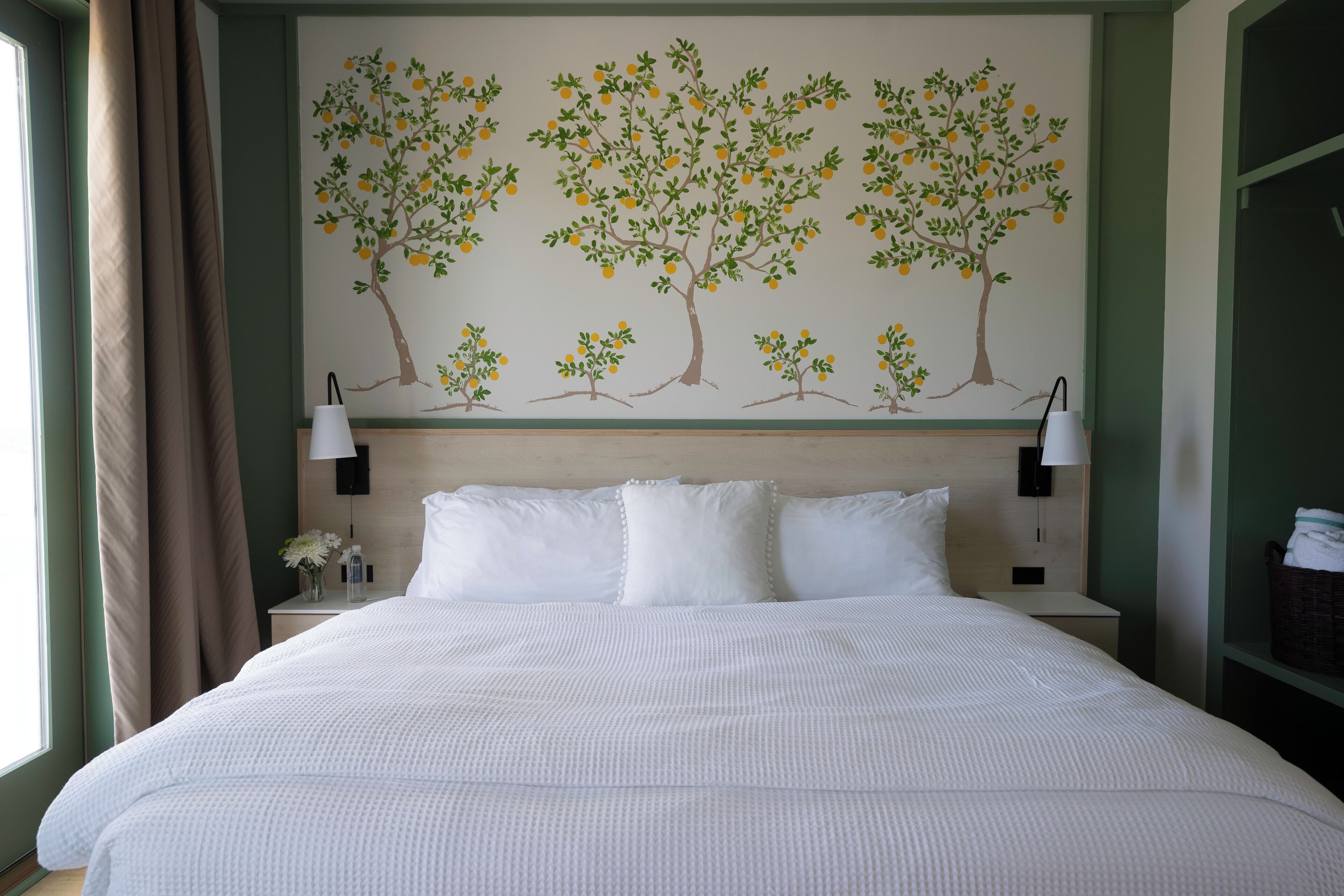 A hotel room with a large bed, two bedside lamps, a nightstand with a vase of flowers, and a wall mural of trees.