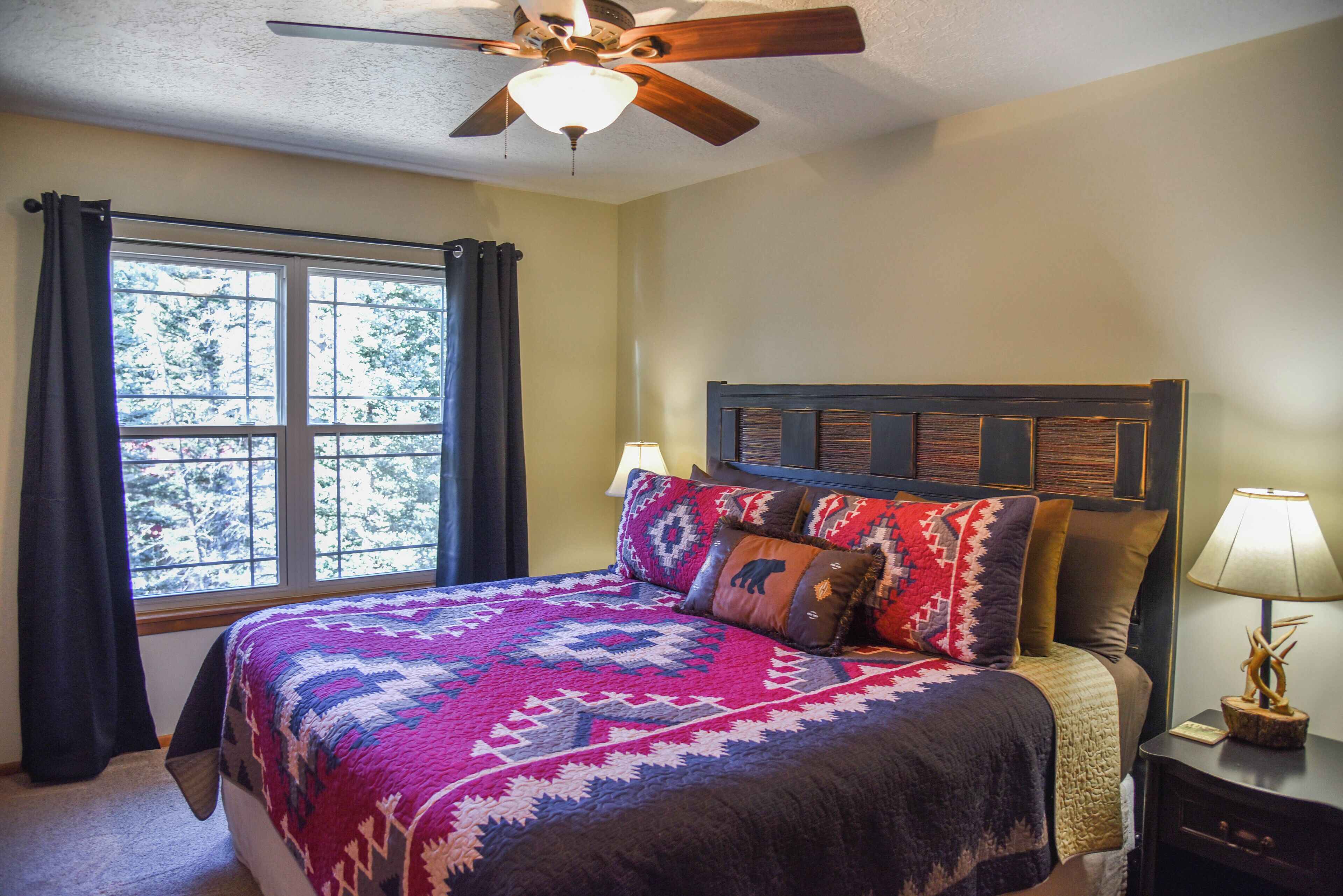 A bedroom with a bed, a ceiling fan, a window with curtains, and a nightstand with a lamp.