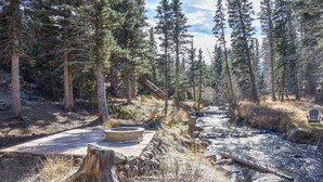 Un cadre naturel en plein air avec un foyer en pierre, un banc en bois et une chaise près d’un ruisseau, le tout entouré d’arbres.