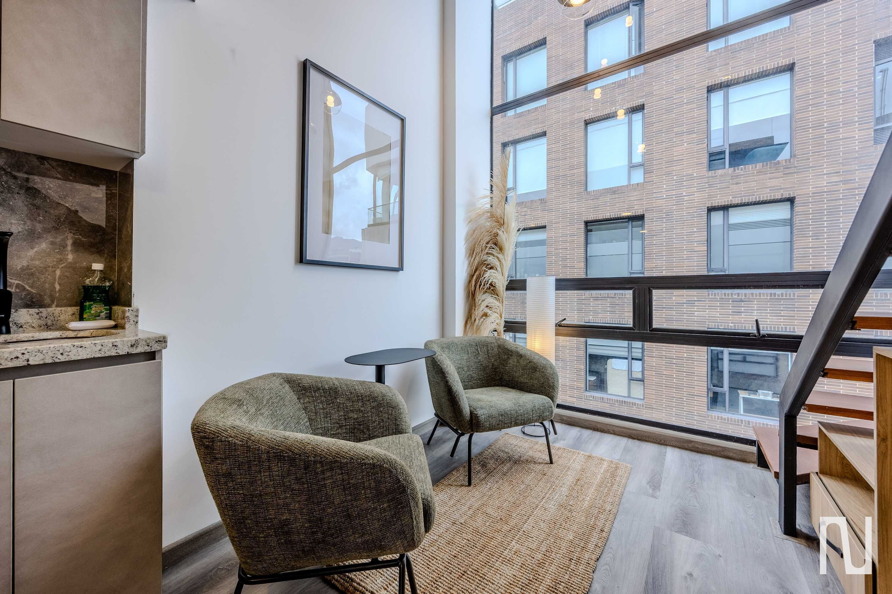 Een modern interieur met twee fauteuils, een klein rond tafeltje, een ingelijste afbeelding en uitzicht op een bakstenen gebouw door grote ramen.
