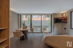 Una habitación moderna con cama, una mesa redonda con sillas, televisor y ventanas amplias.