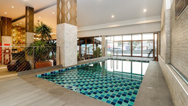 An indoor swimming pool with a tiled floor, surrounded by a wooden deck and a seating area with chairs and potted plants. - Ja Siam City (Pattaya)