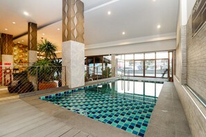 An indoor swimming pool with a tiled floor, surrounded by a wooden deck and a seating area with chairs and potted plants. - Ja Siam City (Pattaya)