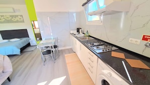 A compact kitchen with a gas stove, a microwave, and a washing machine. There is a dining area with a table and chairs, and a bed with a headboard. - Studio Apartment (Santana)