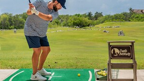 Een persoon speelt golf op een groene mat met een bord waarop 'Puerto Aventuras' staat.