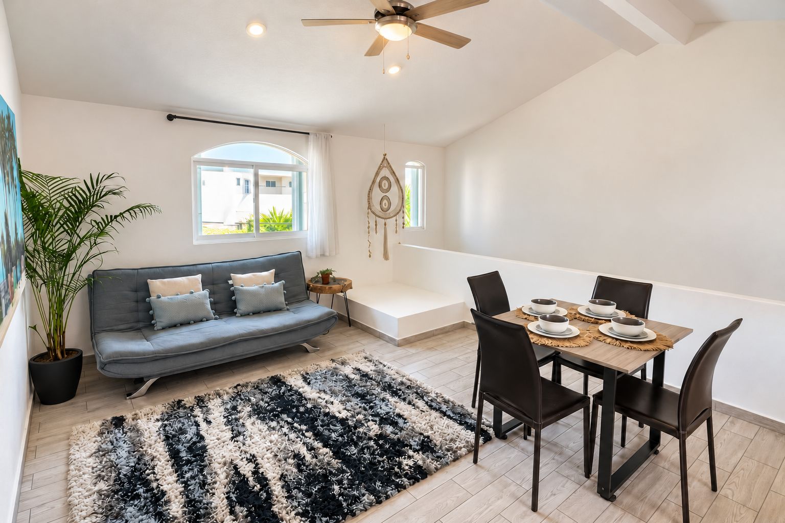 A modern living room with a sofa, dining table, chairs, a ceiling fan, and a large window.