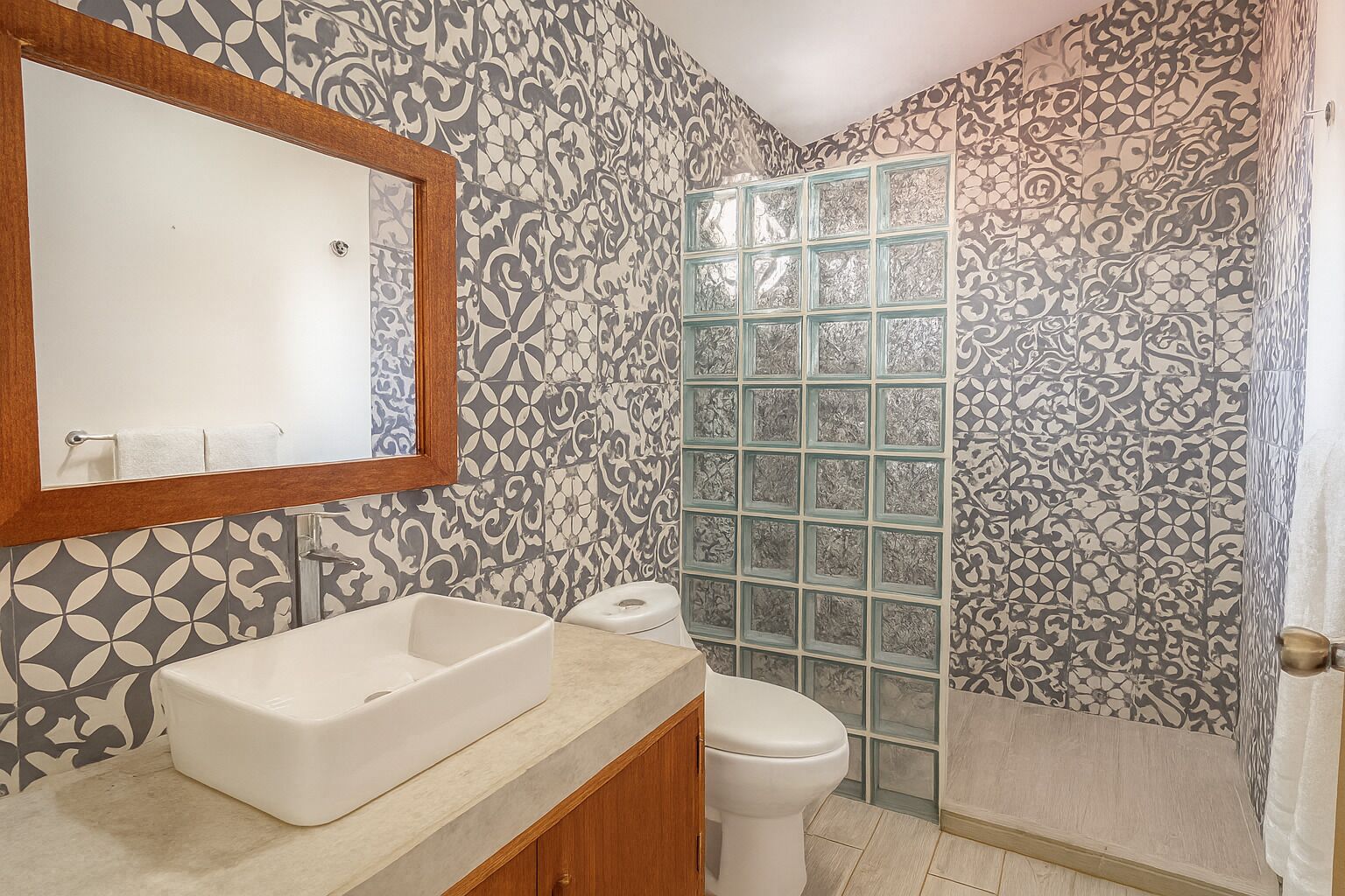 Ein Badezimmer mit großem Spiegel, weißem Waschbecken, Toilette und Dusche mit gemusterten Fliesen.