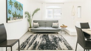 Een moderne woonkamer met een grijze bank, een rond bijzettafeltje, een eethoek met een tafel en stoelen, een groot schilderij van palmbomen en een potplant.