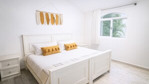 Een witte slaapkamer met een tweepersoonsbed, twee nachtkastjes, een raam met uitzicht op groen en een hangende decoratie.