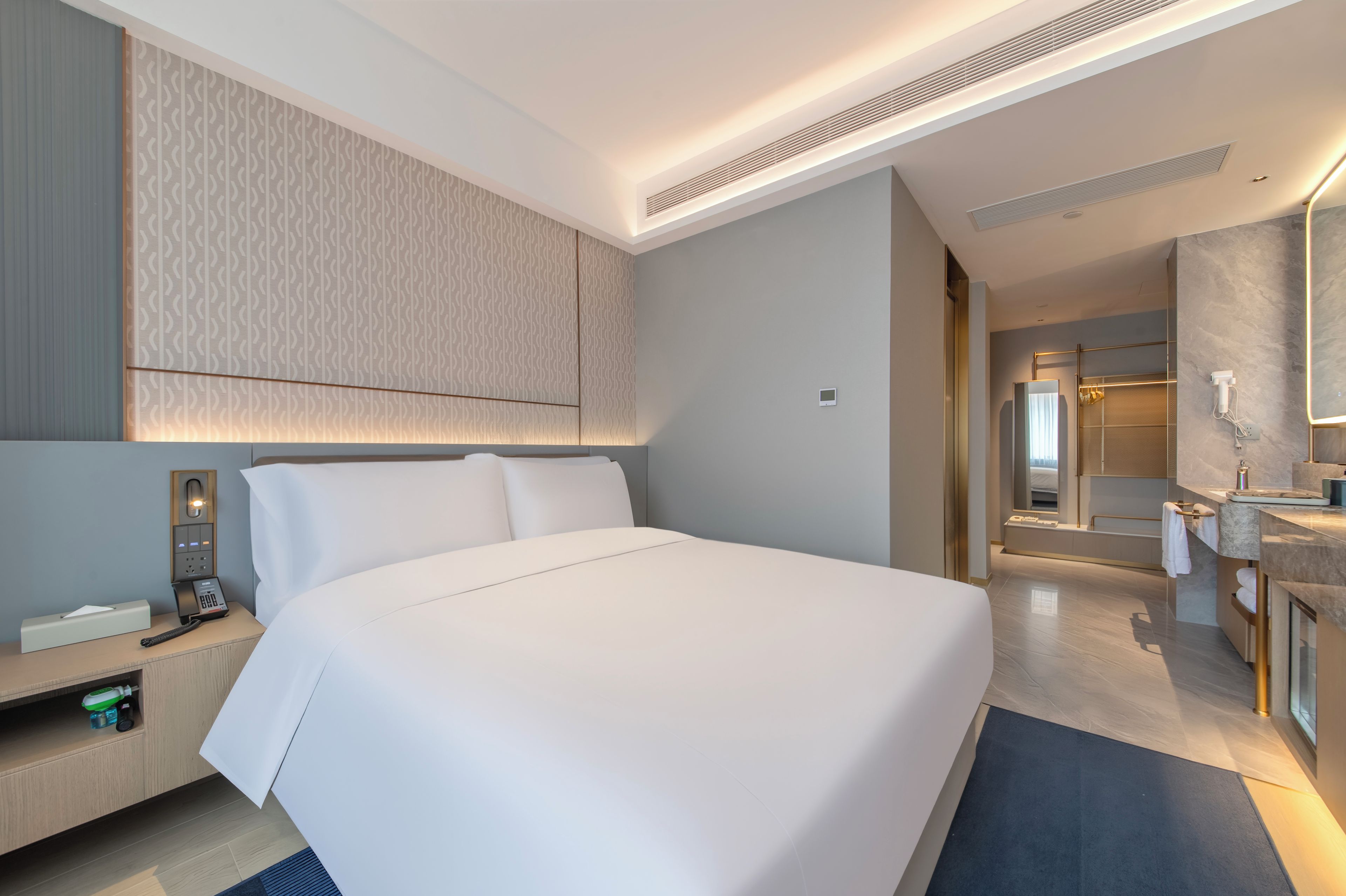Quarto de hotel moderno com uma cama grande, uma mesa de cabeceira com telefone e lenços, parede estampada e banheiro com espelho e pia.