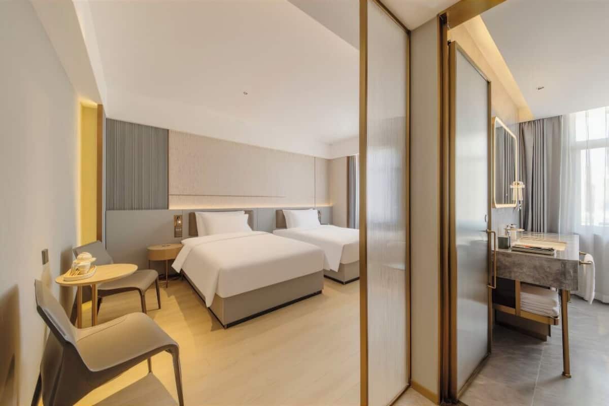 A hotel room with two beds, a desk, a chair, a small table with a cup, and a mirror.