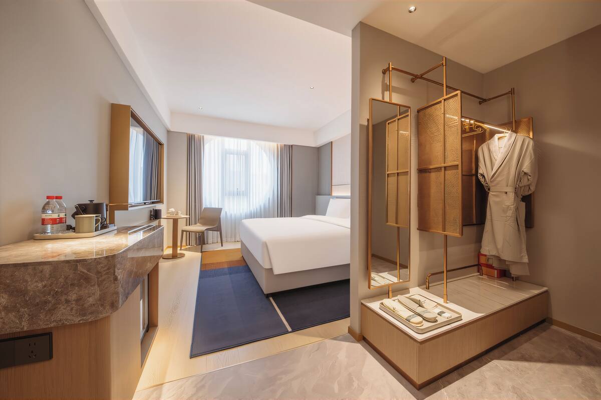 A modern hotel room with a bed, a vanity with a mirror, a robe hanging, and a desk with a chair.
