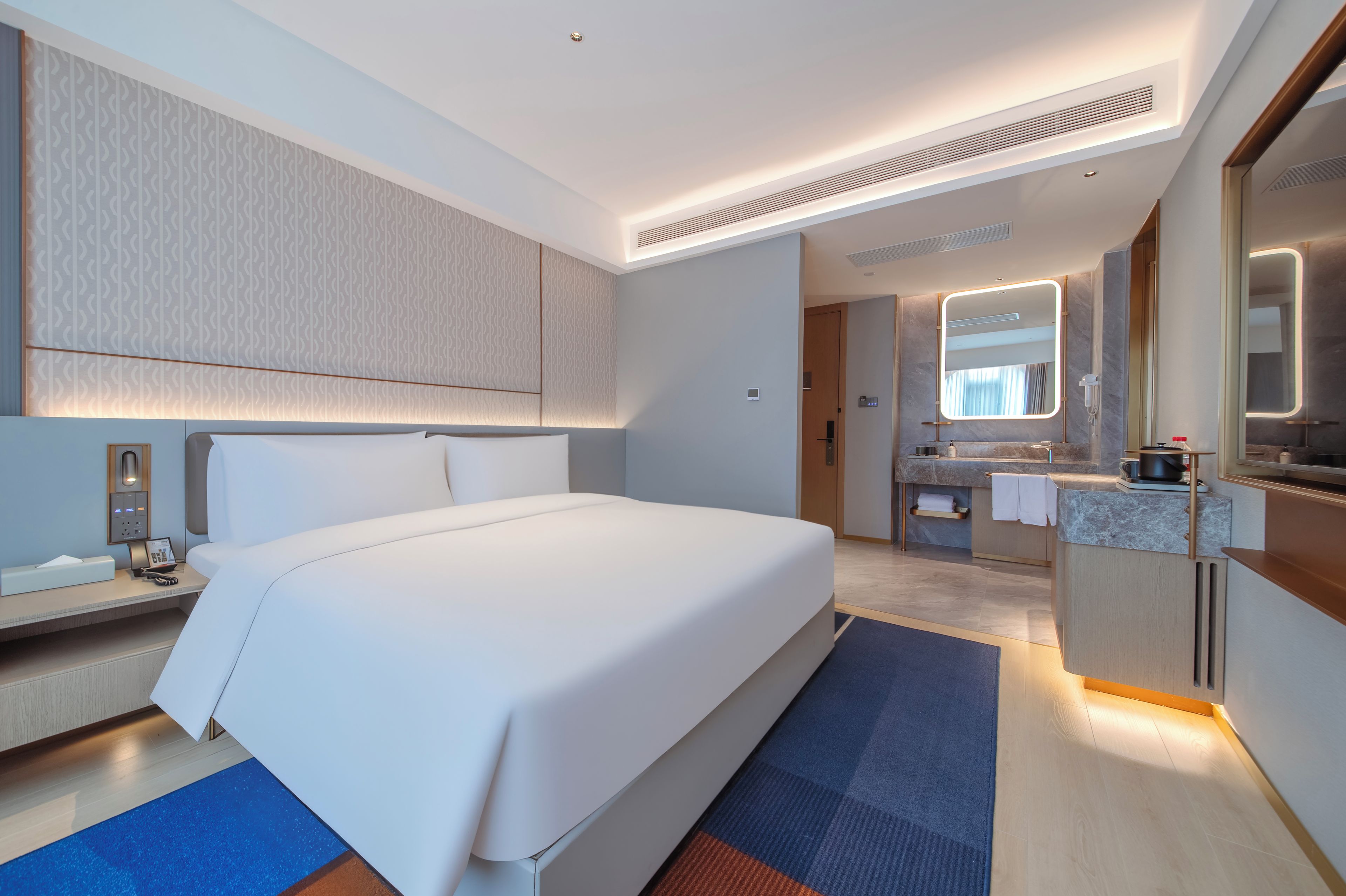 Una habitación de hotel moderna con una cama grande, un baño con tocador y espejo, y una pequeña cocina.