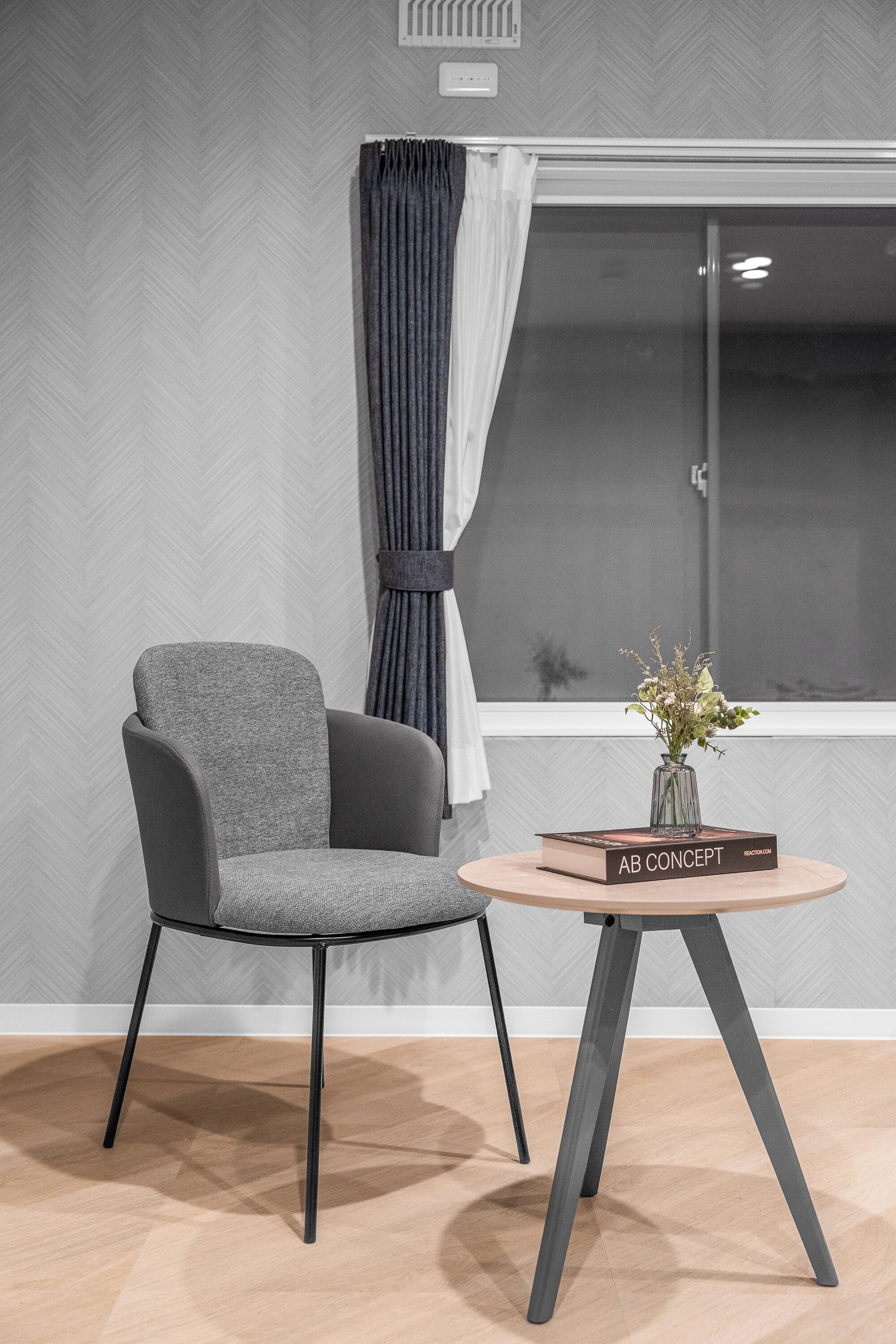 Une pièce moderne avec un fauteuil à revêtement gris, une table ronde en bois sur laquelle est posé un vase, et une fenêtre avec des rideaux.