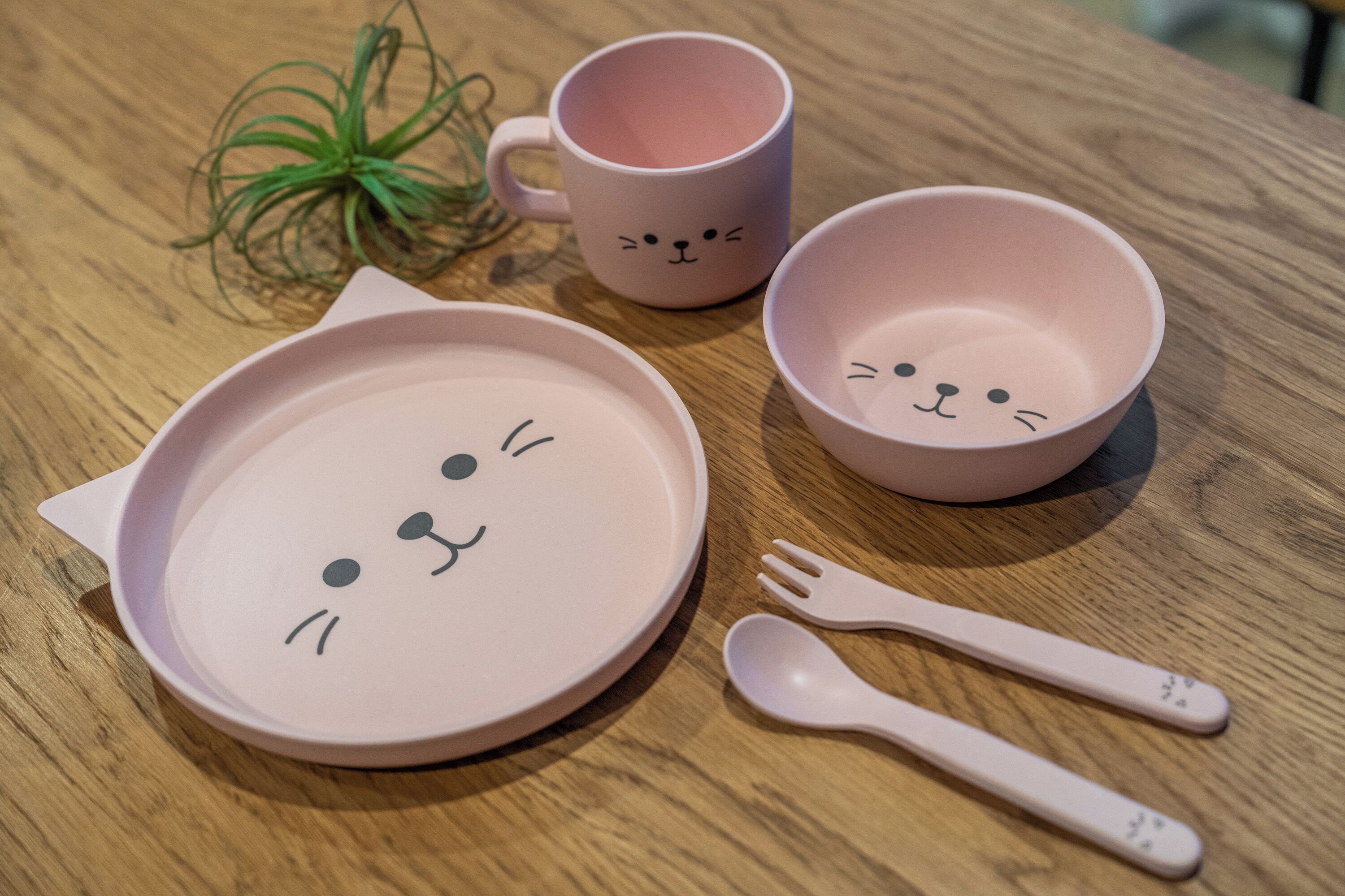 A table with a pink cat-themed plate, a pink mug, a pink bowl, and a pink spoon.