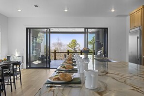 Une cuisine moderne avec une longue table à manger dressée pour le repas, un réfrigérateur et une vue dégagée grâce à de grandes portes-fenêtres coulissantes.