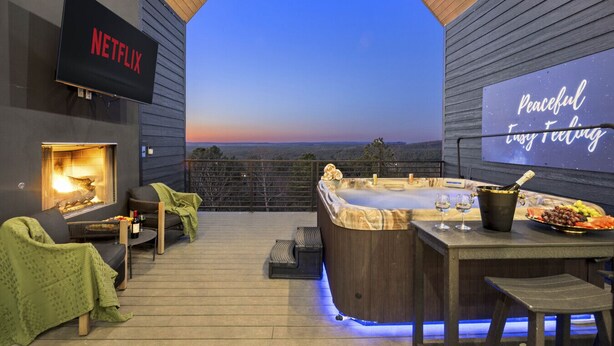 A cozy outdoor area with a fireplace, seating, a hot tub, and a TV screen displaying 'NETFLIX'. - Eagle`s Vista · Heated Pool, Stunning Views, Hot Tub & Game Room - Sleeps 30! (Broken Bow)