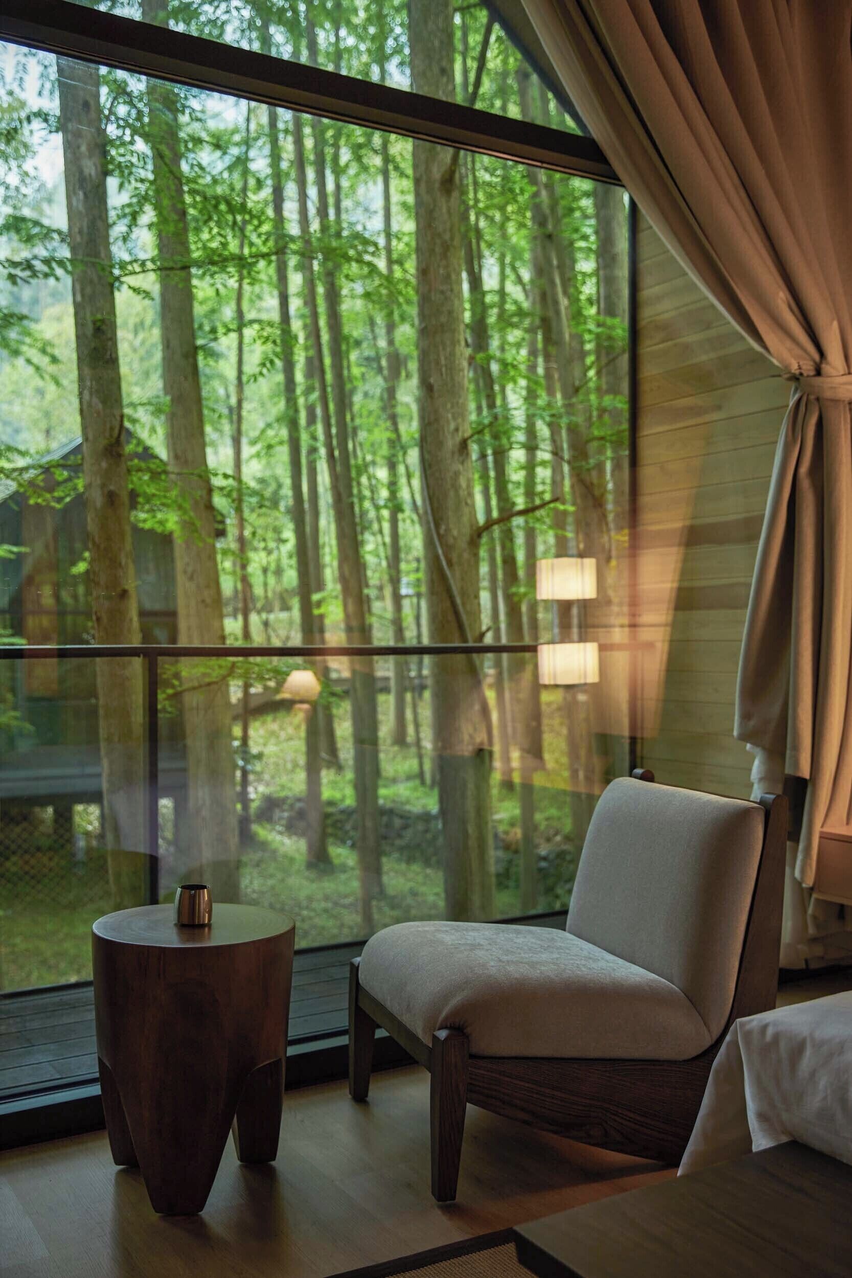 A room with a large window overlooking a forest, a chair, a small round table, and a lamp.