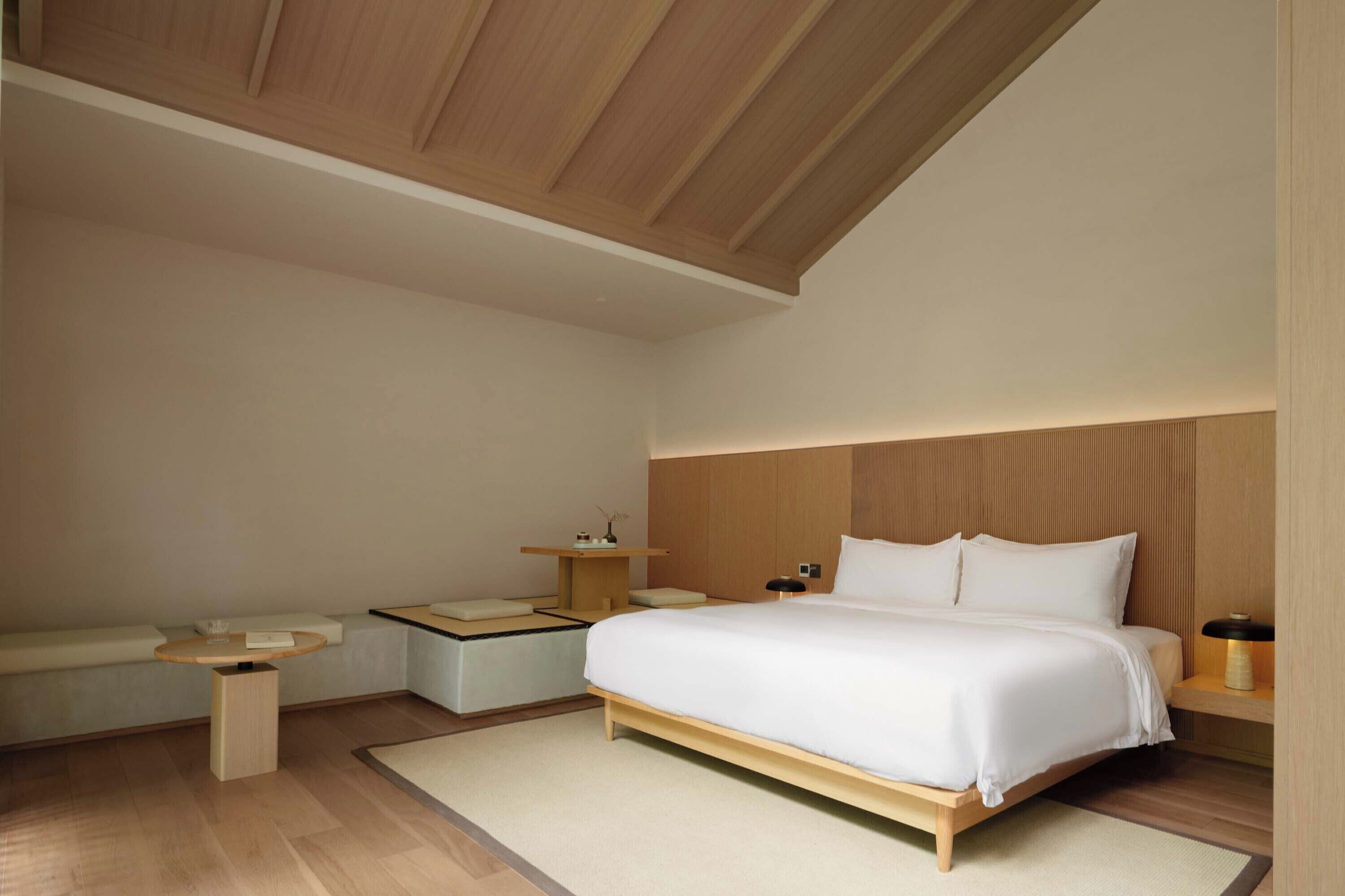 A modern bedroom with a large bed, a bedside table, a small table, and a bench.