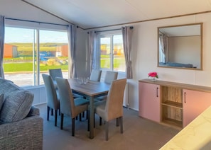 A room with a dining table, chairs, a sofa, a cabinet, and a large window with curtains. - Pondview Retreat Lodge (Pet) (Glenluce, Newton Stewart)