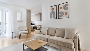 Un soggiorno moderno con un divano beige, un tavolino in legno e una poltrona bianca. Si nota una cucina sullo sfondo con mobili bianchi e un forno a microonde. Due opere d'arte incorniciate adornano la parete.