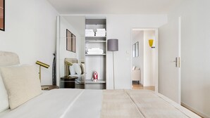 Una camera da letto con un letto, un comodino con una lampada, un armadio con ripiani e una porta che dà su un'altra stanza.