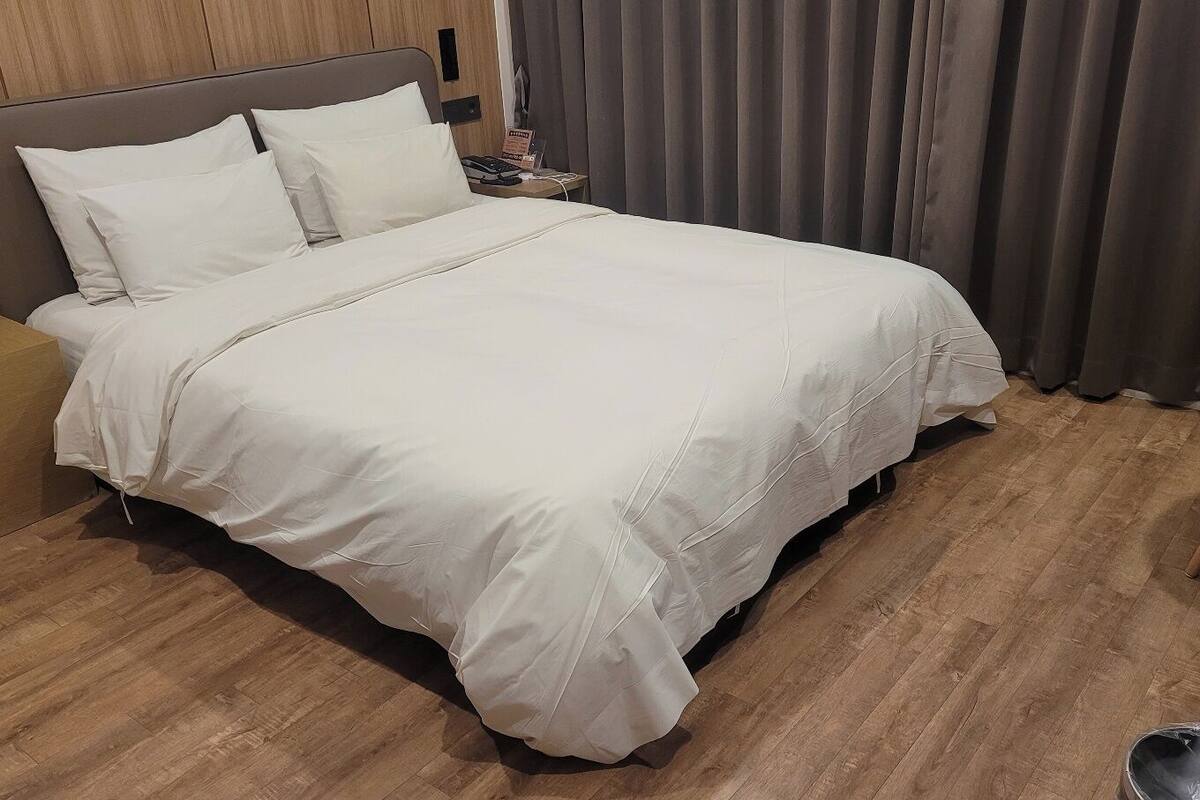 A neatly made bed with white bedding, a wooden headboard, a nightstand with a phone, and a chair with a white table.