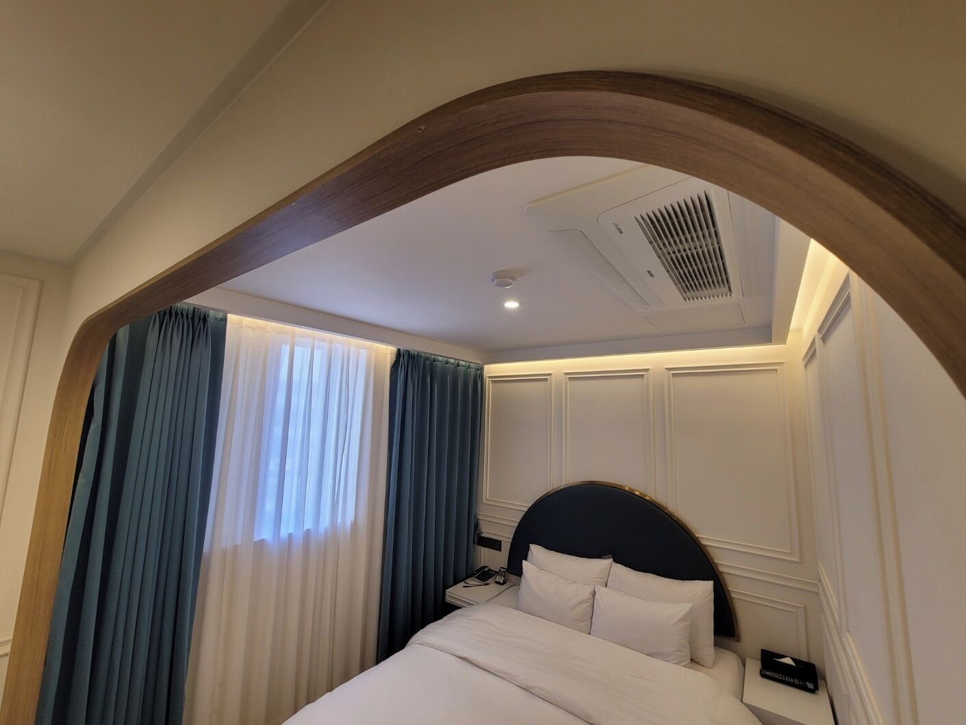 A hotel room with a curved wooden arch, a bed with white linens, a nightstand, and a window with blue curtains.