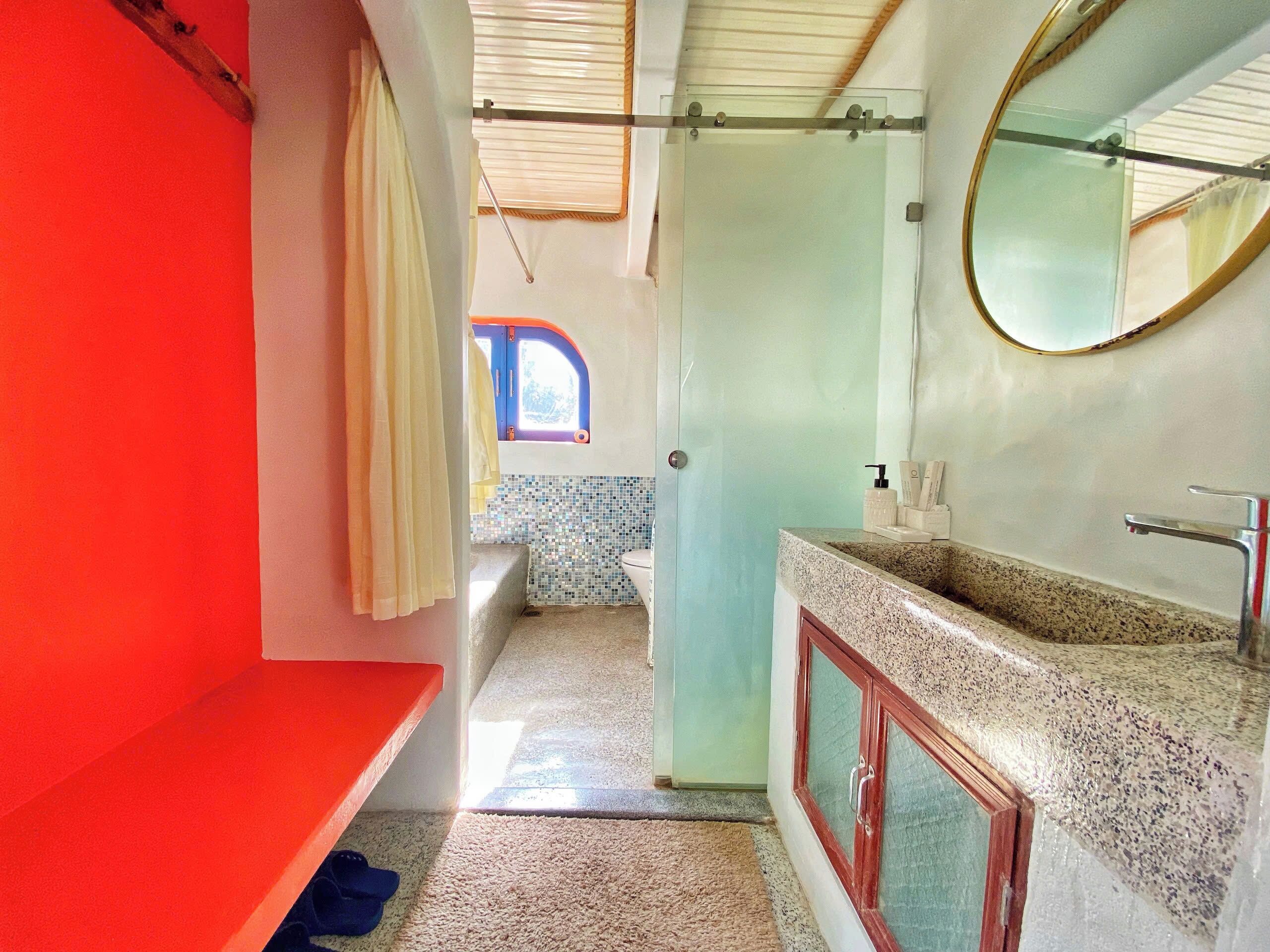 A bathroom with a red wall, a glass shower enclosure, a sink with a mirror, and a small window.