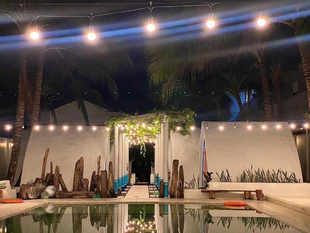 Un área de piscina al aire libre con un sendero que lleva a una zona de estar cubierta, rodeada de palmeras e iluminación decorativa.