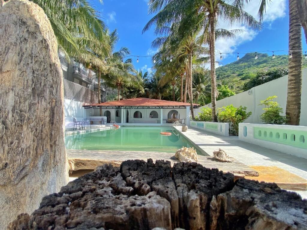 Ein Poolbereich mit einem Gebäude im Hintergrund, umgeben von Palmen und einem Berg.