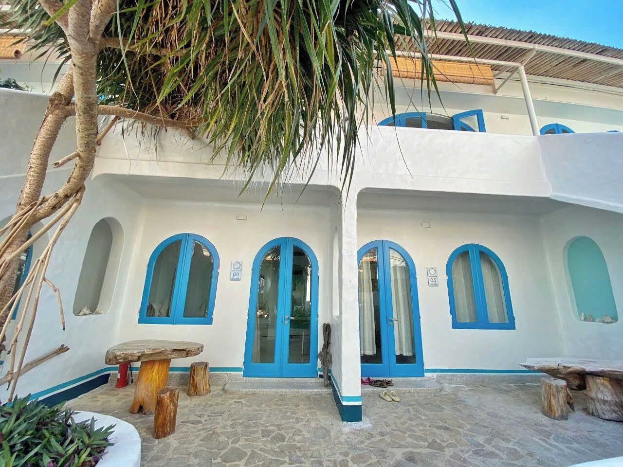 A building with white walls and blue accents, featuring arched windows and doors, a stone courtyard, and rustic wooden furniture.